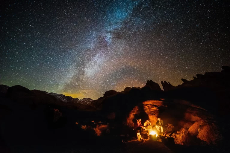 A group of people gathered around a bonfire with mountains in the background.