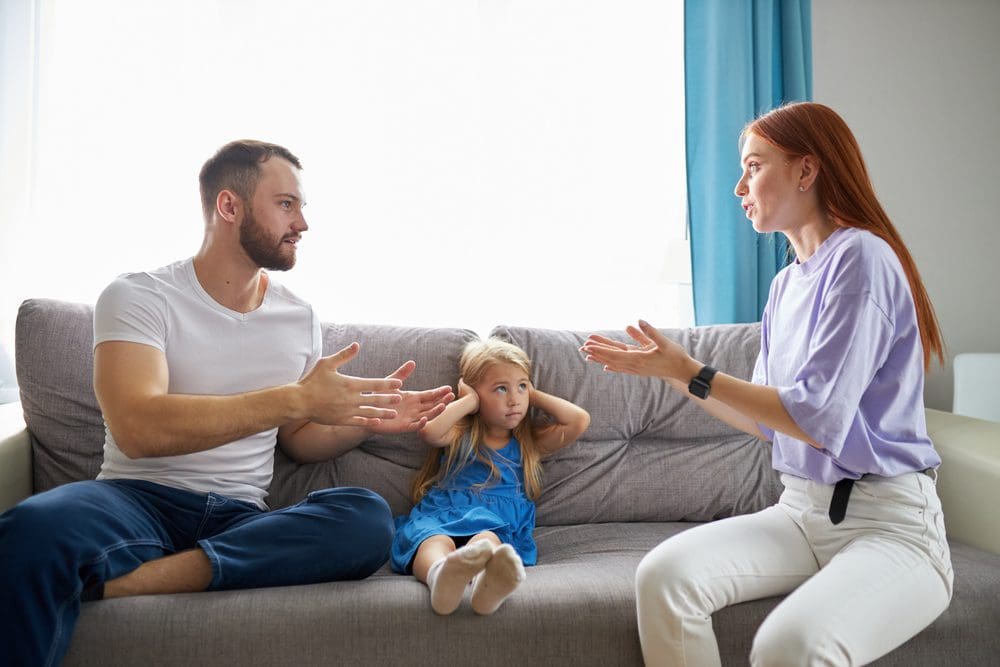 Parents arguing during a custody exchange while their daughter looks distressed, highlighting the emotional toll of co-parenting conflicts.