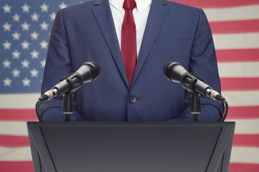 Man in a suit standing at a podium with microphones, American flag in the background.