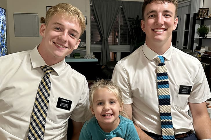 Two young male LDS missionaries, dressed in white shirts and ties, smile warmly while sitting next to a young child in a living room setting
