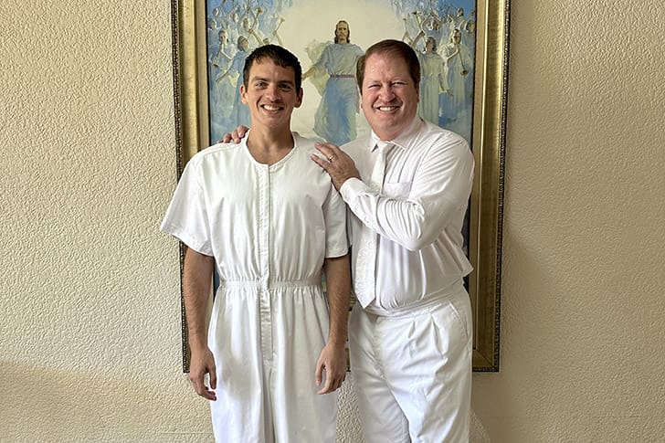  A newly baptized member of the Church, dressed in white, stands next to smiling Scot in front of a painting depicting the Savior, Jesus Christ.