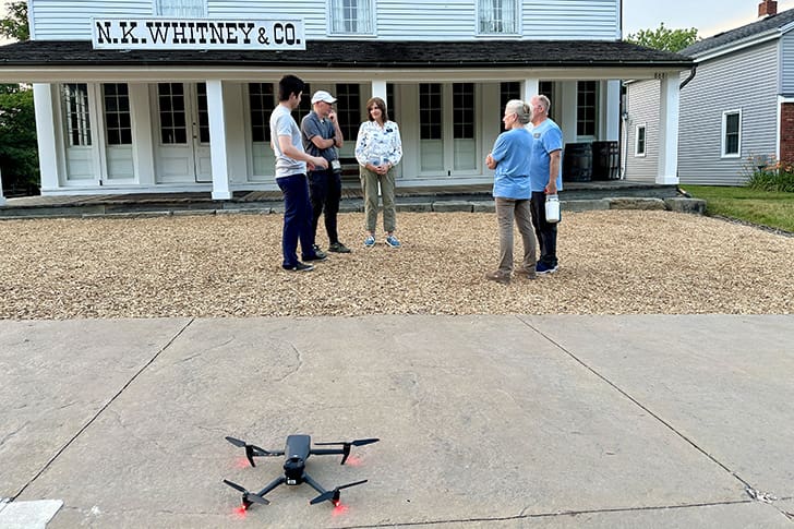 A group of people, including Maurine, stands outside the historic N.K. Whitney & Co. building in Kirtland, Ohio, with a drone on the ground, capturing the scene.