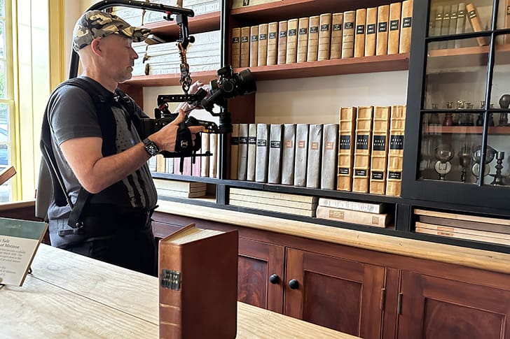 A videographer films inside a historic bookstore, capturing the essence of early LDS Church history with shelves of first editions of Books of Mormon in the background.