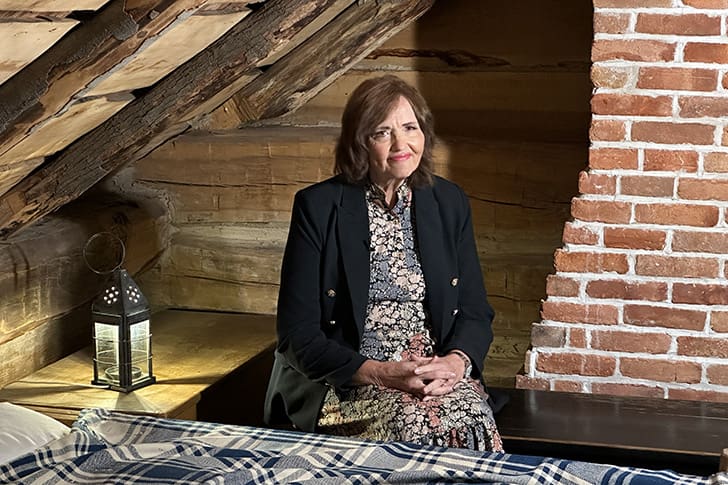 Muarine sits in a cozy, rustic room under a sloped roof, with exposed brickwork and wooden beams, prepared to discuss the marvelous event that took place in this holy airspace. 