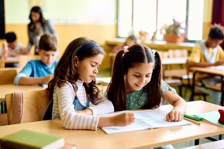 Two young girls sharing a book in a classroom, learning and making new friends.