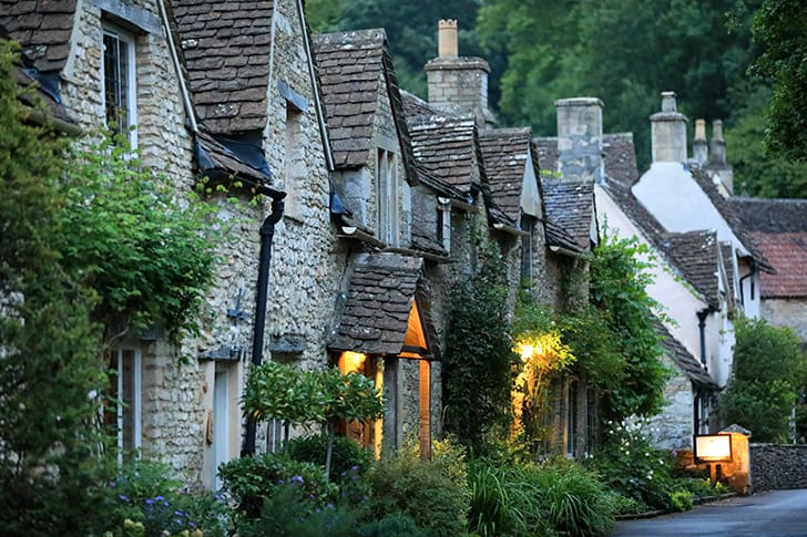 n evening view of Castle Combe’s stone cottages, softly illuminated by warm lights, highlighting the village’s serene atmosphere.