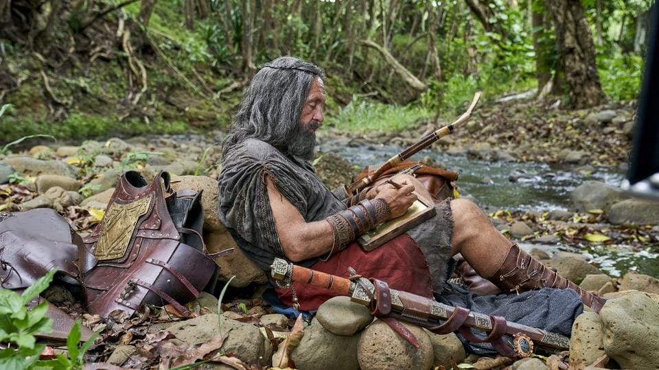 Moroni writing in the Book of Mormon by a stream, with armor and weapons nearby, representing his dual role as a prophet and defender of faith.