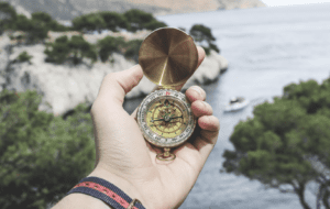 Hand holding a brass compass near a coastline, representing clarity, purpose, and navigating challenges through True North leadership principles.