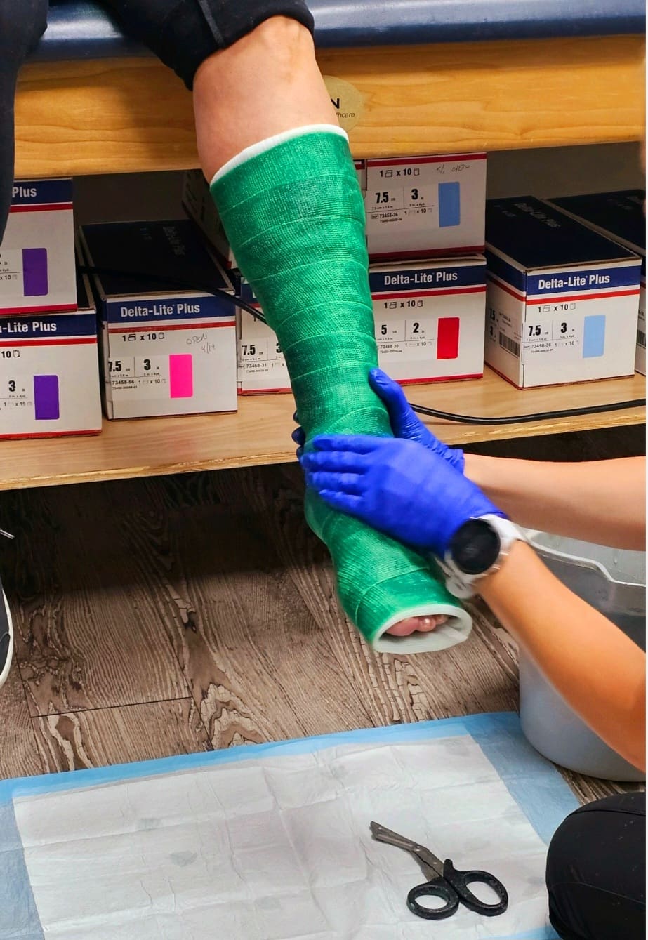 A medical technician applies a green orthopedic cast to a patient’s lower leg, symbolizing the beginning of a journey of healing, patience, and gratitude