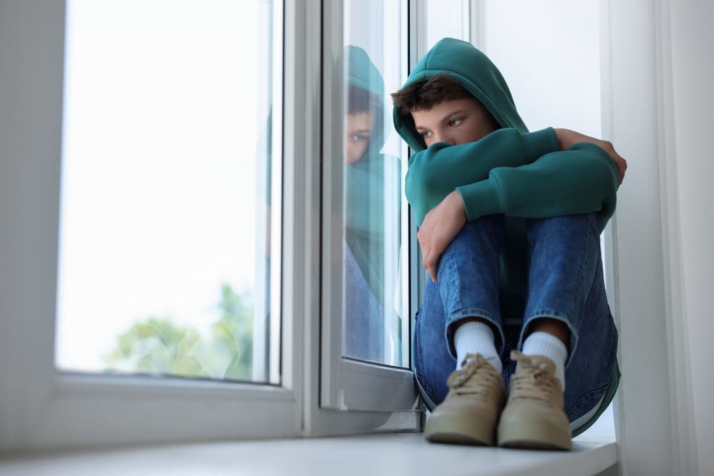Teen boy in hoodie sitting by window, showing signs of depression and emotional withdrawal