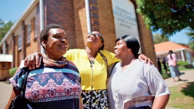 Three Latter-day Saint women smiling outside a Church meetinghouse, representing global unity and gospel-centered community.