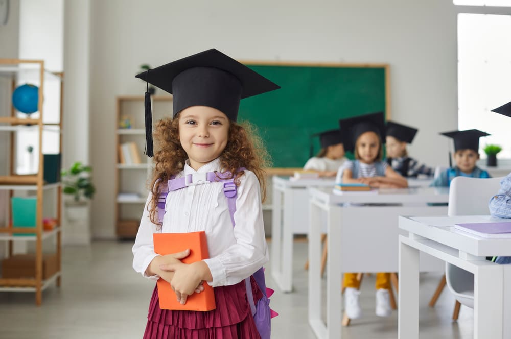 Young girl in graduation cap holding a book in a classroom, celebrating school milestones and the joy of learning.
