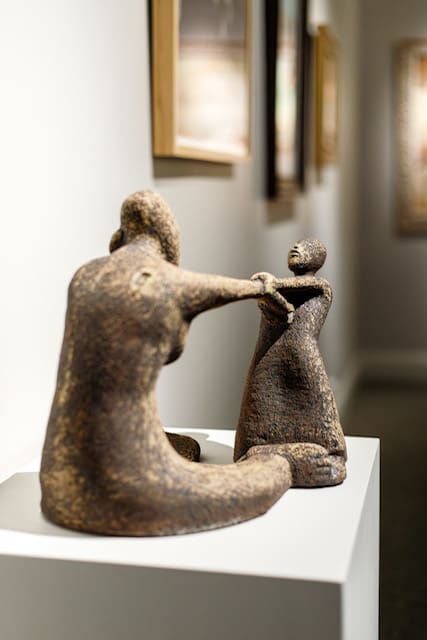 A clay sculpture featured at the 13th International Art Competition shows one figure reaching toward another in a gesture of compassion and support, exemplifying the theme of “succor the weak, lift up the hands which hang down.” This artwork visualizes the divine directive from Doctrine and Covenants 81:5.