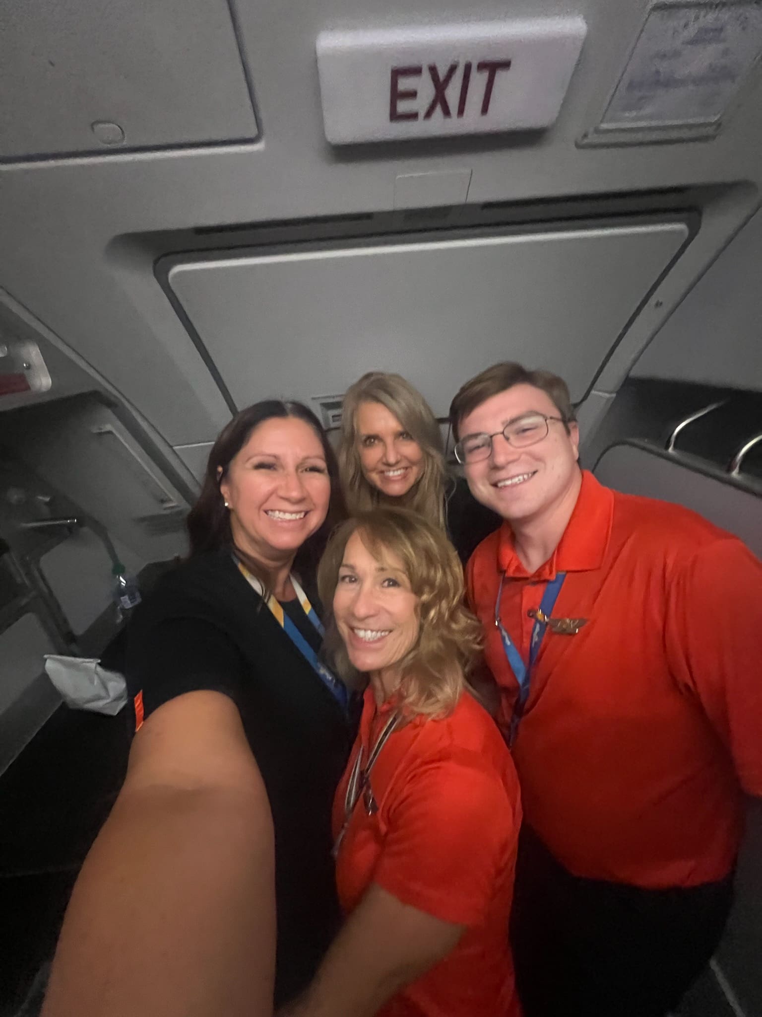 Four Allegiant flight attendants and crew smile for a selfie in front of the exit row door during a late-night flight, representing their close-knit bond in the skies.