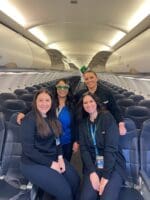 Four flight attendants pose together in an empty Allegiant airplane cabin before takeoff, symbolizing their preparation to serve passengers with love and faith.