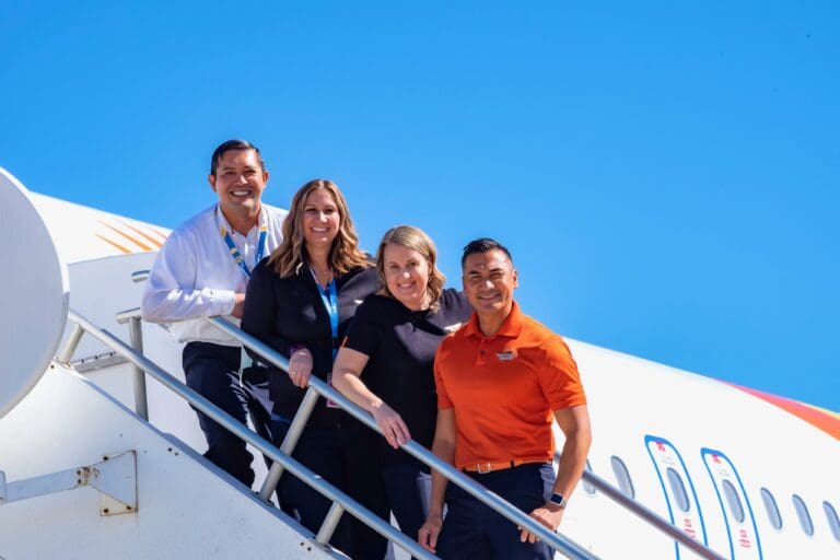 Allegiant flight attendants, including Holly Houskeeper, smiling on airplane stairs under blue sky as flight attendant shares gospel during service.