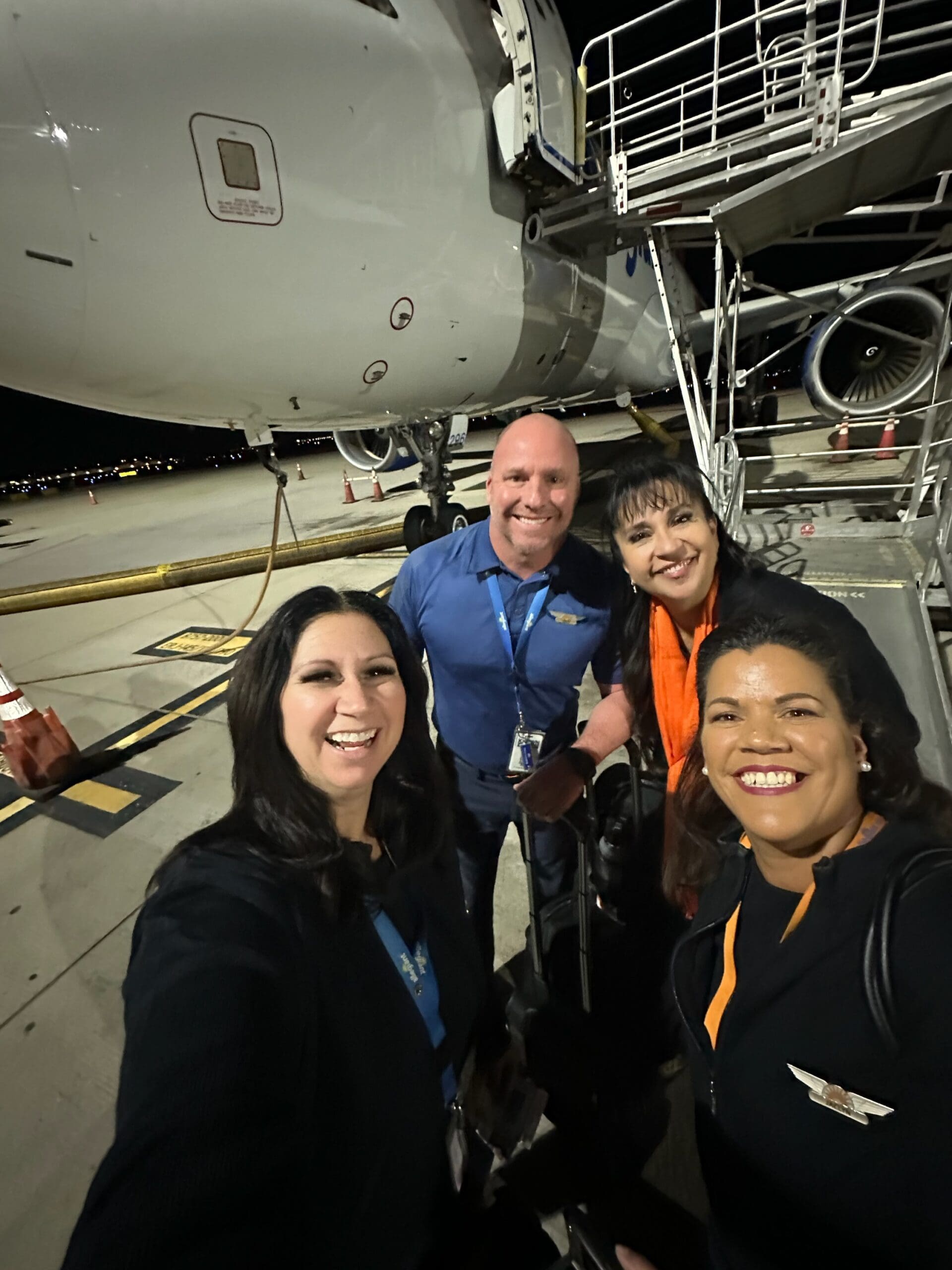 A group of Allegiant flight attendants smiles together on the tarmac at night, standing near the aircraft stairs after a completed flight, reflecting their unity and support.