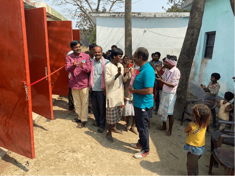 A group of villagers in India celebrates the ribbon-cutting ceremony for newly built toilets and showers in a leprosy colony. The facilities were provided as part of a service initiative led by Becky Douglas and her humanitarian organization, emphasizing humility, clean water access, and disease prevention