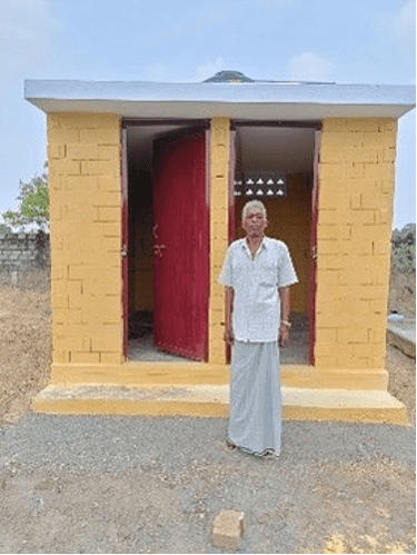 A man stands proudly in front of a newly constructed toilet facility built through service efforts to improve sanitation and restore dignity in an Indian village.