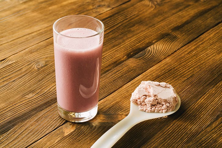  A glass of pink meal replacement shake with protein powder on a spoon, illustrating the difference between balanced shakes and high-sugar hospital options