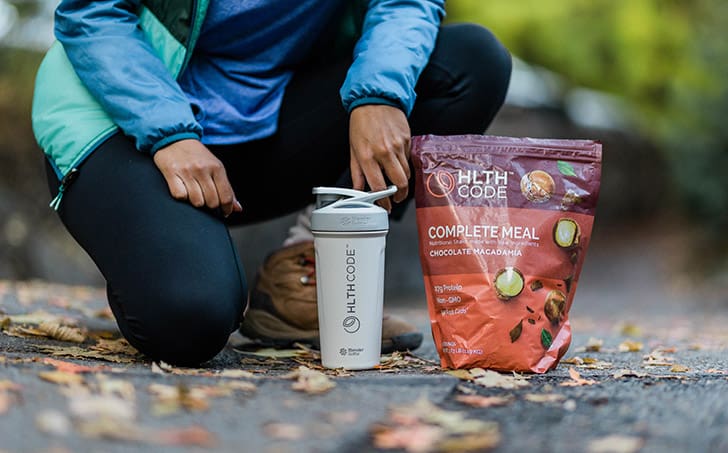 Person kneeling outdoors beside a bag of HLTH Code Complete Meal and shaker bottle, showcasing convenient nutrition for weight loss and metabolism support.