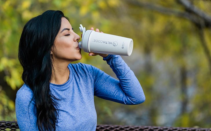 Woman enjoying HLTH Code Complete Meal shake outdoors, demonstrating convenient meal replacement for healthy weight management and energy.