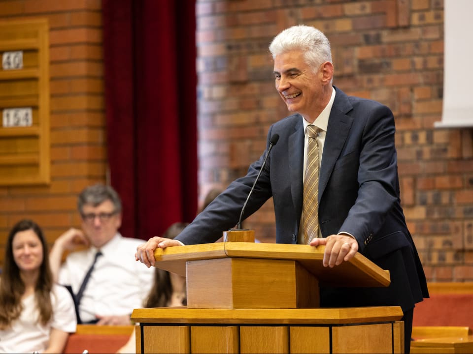 Elder Gérald Caussé, newly called to the Quorum of the Twelve Apostles, smiles as he speaks at a Latter-day Saint meetinghouse pulpit. Known for his warm leadership and emphasis on ministering, the former Presiding Bishop continues to inspire members worldwide with his testimony of Jesus Christ and commitment to global Church service.