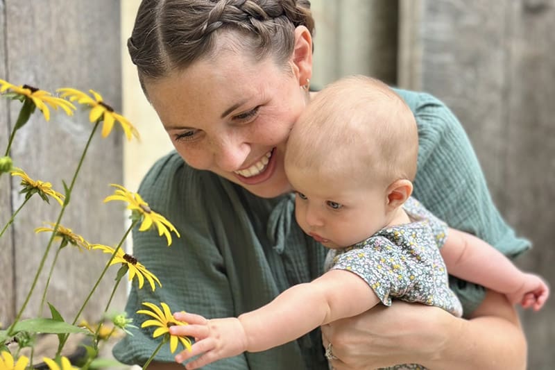 Michaela Proctor Hutchins, host of the Measure of Her Creation podcast, smiles as she holds her baby and helps her reach for yellow flowers. The image captures the spirit of motherhood, divine purpose, and revelation central to the podcast’s message about how Latter-day Saint women balance faith, family, and calling on the covenant path.