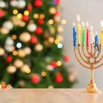 Menorah with colorful candles glowing in front of a decorated Christmas tree, representing Hanukkah traditions inspiring Christmas preparation and family traditions.