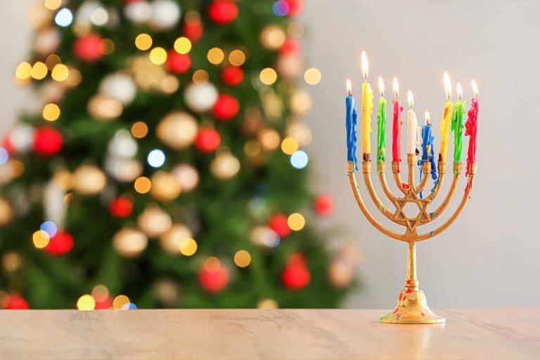 Menorah with colorful candles glowing in front of a decorated Christmas tree, representing Hanukkah traditions inspiring Christmas preparation and family traditions.