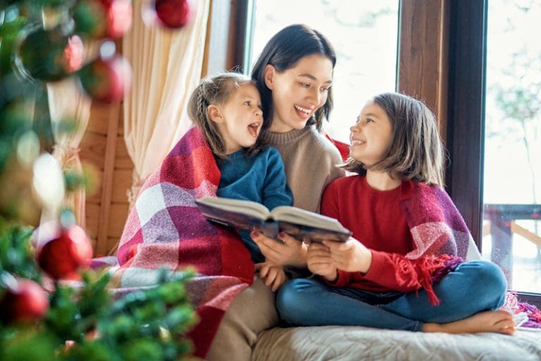 Mother reading with two children by the Christmas tree, representing cozy winter moments inspired by Christmas activity books for kids and creative holiday kits.