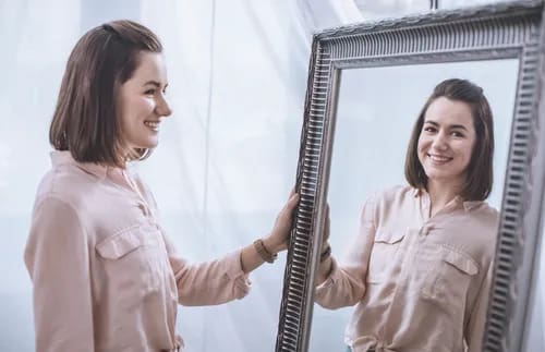 Woman smiling at her reflection, representing confidence gained through faith, persistence, resilience, and steady personal growth.