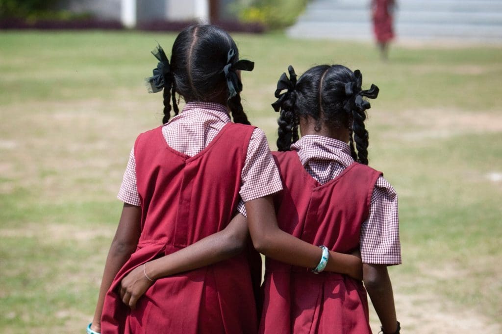 Two school-age girls in matching uniforms walk arm in arm across a grassy field, symbolizing companionship, trust, and lasting friendship.