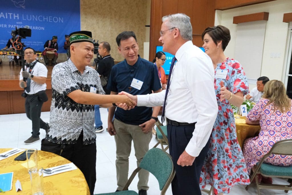 Elder Patrick Kearon of the Quorum of the Twelve Apostles greets attendees at a Church gathering alongside his wife, Jennifer Kearon. The image reflects his global ministry and leadership role within The Church of Jesus Christ of Latter-day Saints as he prepares to address young adults worldwide in a global young adult devotional commemorating the 100-year anniversary of Institutes of Religion.