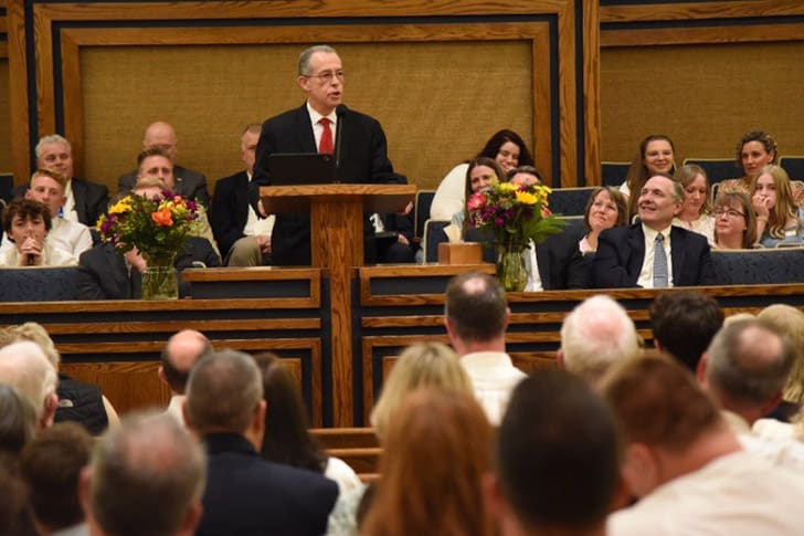 Church leader speaking at a podium to a large audience, symbolizing leadership and teaching within the Church.