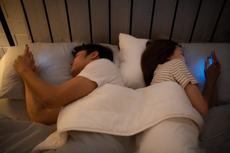 Couple in bed looking at smartphones instead of each other, illustrating digital connectivity replacing real human connections and authentic relationships